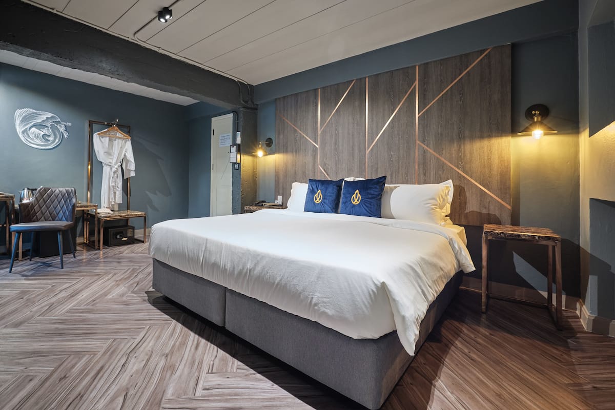 Moderni hotellihuone, jossa on suuri sänky, puinen pääty, oleskelualue tuolilla ja pöydällä sekä kylpytakki henkarissa.