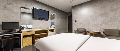 A modern hotel room with a large bed, a desk with a computer, a television, and a seating area with a chair and a small table.