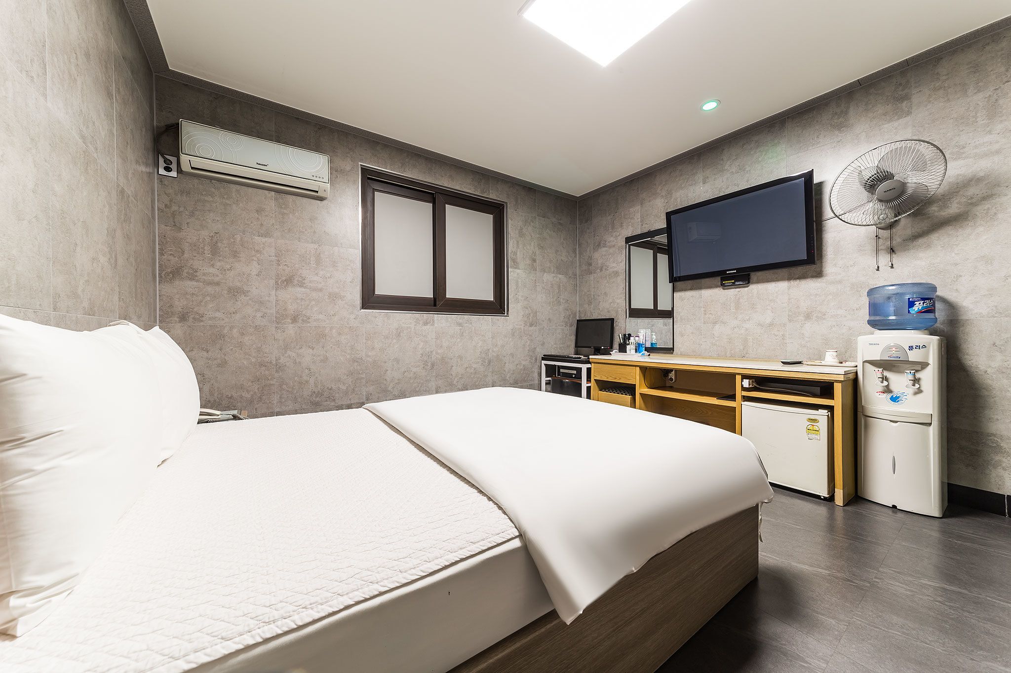 A modern hotel room with a bed, a flat-screen TV, a water dispenser, and a small desk with a computer.