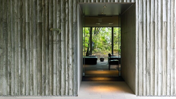 Ein modernes Interieur mit vertikalen Holzpaneelen, einer Sitzecke mit einer Bank und einem Sessel, und Blick auf einen Wald durch große Fenster.