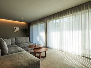 Una sala de estar moderna con un sofá seccional, dos mesas auxiliares de madera y cortinas translúcidas.