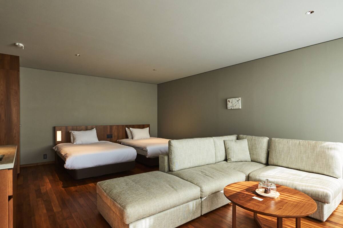 A modern hotel room with a wooden floor, a large L-shaped sofa, two single beds, and a small round wooden table.