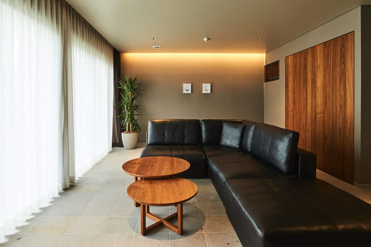 A modern living room with a black leather sofa, two wooden side tables, a potted plant, and a large window with curtains.