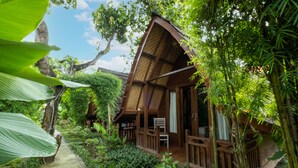 A wooden cabin with a sloped roof, surrounded by lush greenery and a concrete pathway. - Kubu Kak Dudung Villas (Jimbaran)