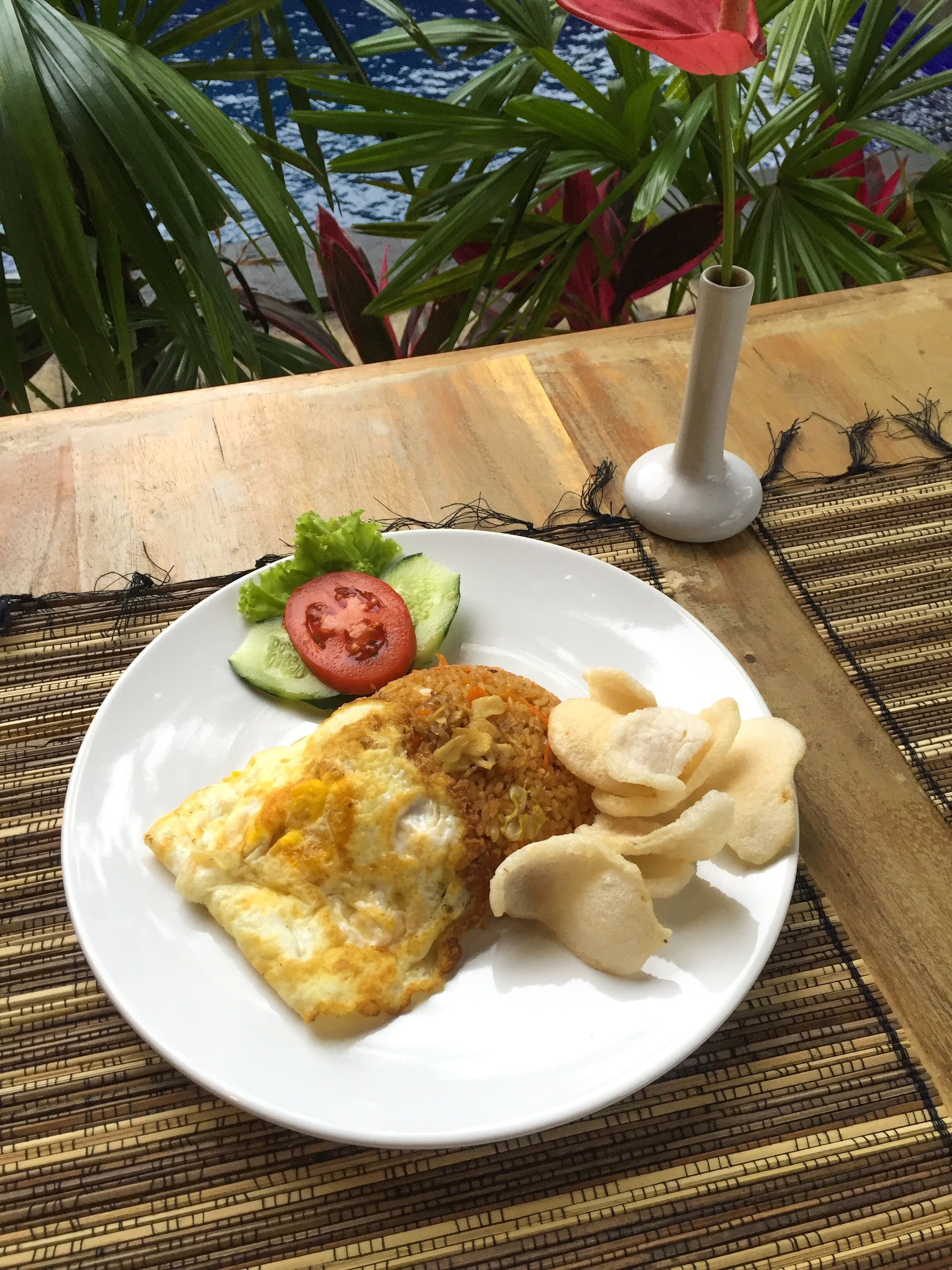 Un plato con arroz frito, un huevo frito y papas fritas, acompañado de una ensalada de lechuga, tomate y pepino, sobre una mesa de madera con un camino de mesa tejido.