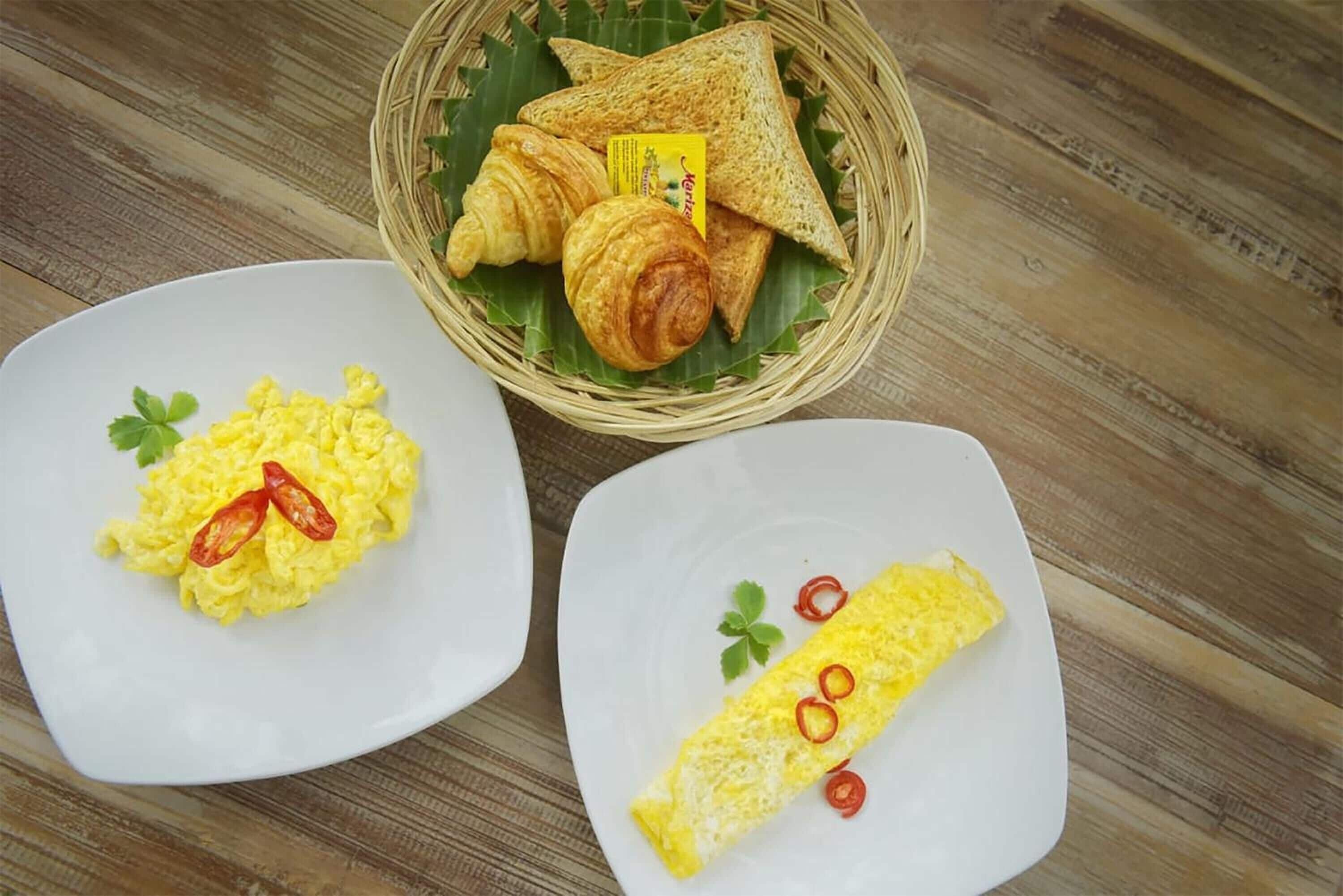 Twee borden roerei en een omelet, met een mand met brood en croissants op de achtergrond.