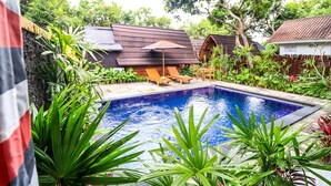 A swimming pool surrounded by lush greenery, wooden lounge chairs, and a small wooden structure with a tiled roof. - Kubu Kak Dudung Villas (Jimbaran)
