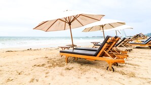 A beach with wooden lounge chairs and beige umbrellas. - Kubu Kak Dudung Villas (Jimbaran)
