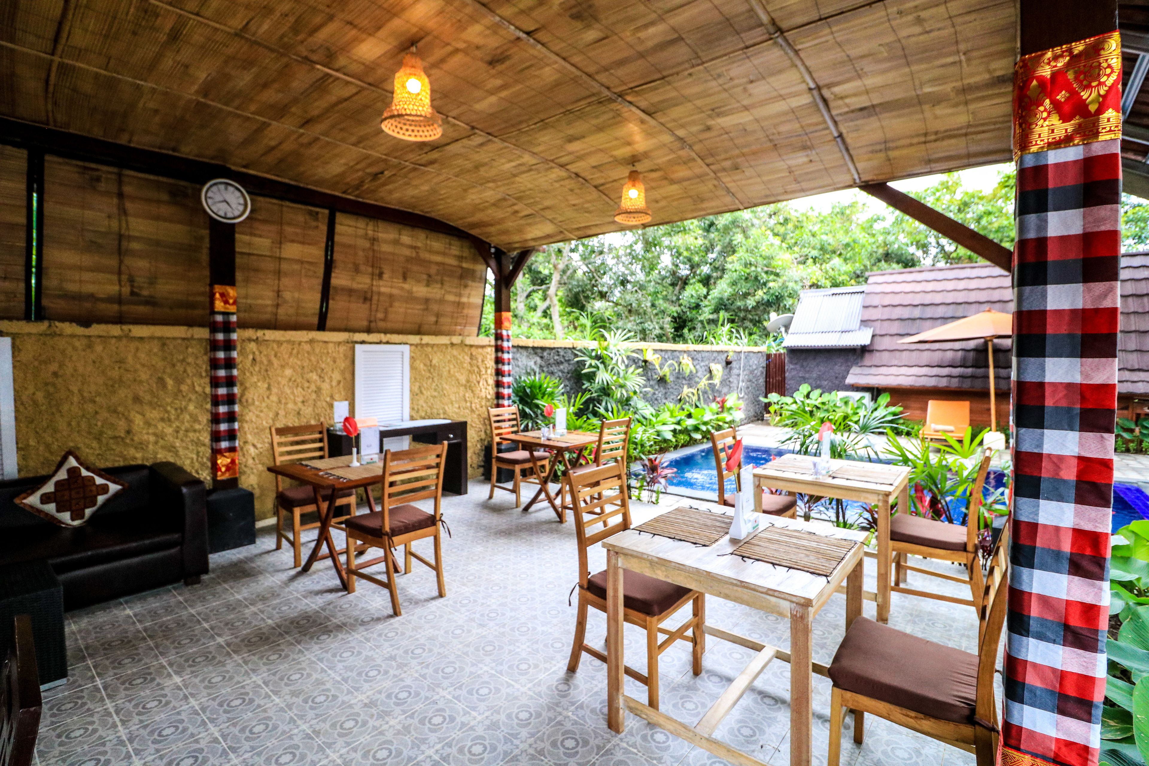 An outdoor dining area with wooden tables and chairs, a clock on the wall, and a checkered cloth hanging on the right.