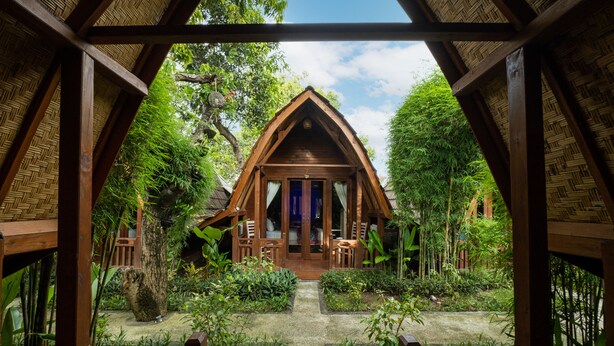 A wooden cabin with a thatched roof, surrounded by lush greenery and a stone pathway leading to the entrance. - Kubu Kak Dudung Villas (Jimbaran)