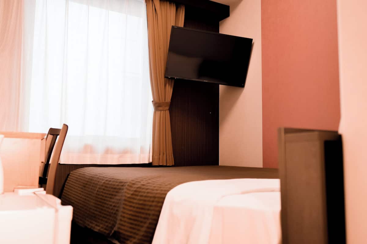 A hotel room with a bed, a chair, a TV mounted on the wall, and a window with curtains.