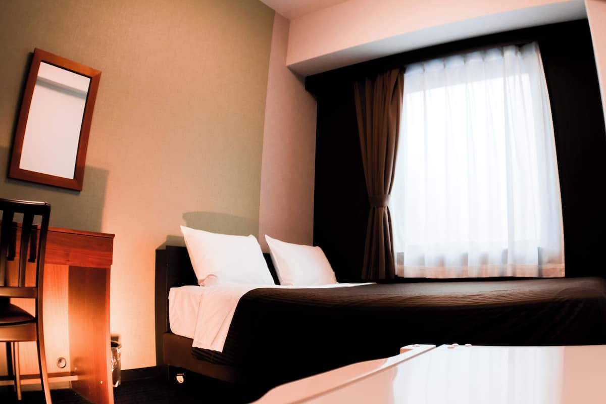 A hotel room with a bed, a chair, a small table, and a window with curtains.