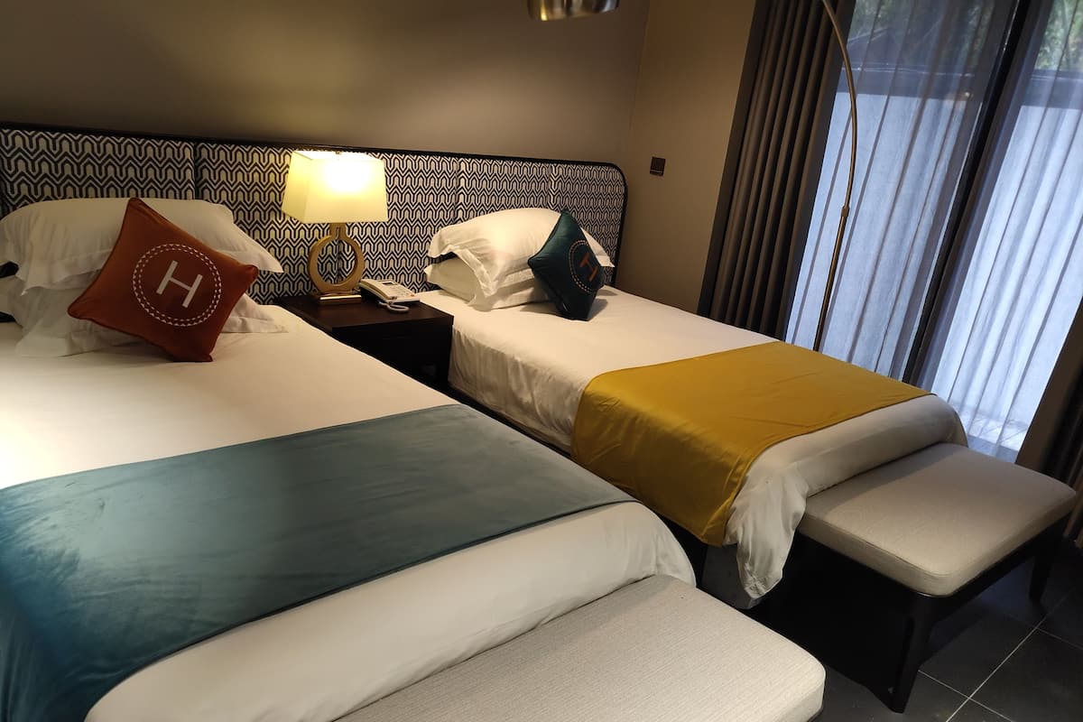 A hotel room with two beds, each with a different colored bedspread, a bedside table with a lamp, and a window with curtains.