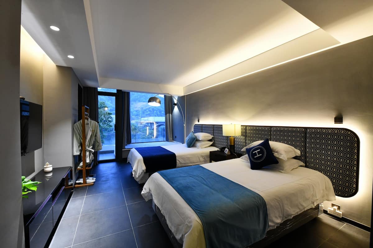 A modern hotel room with two beds, a large window, and a view of the outdoors.
