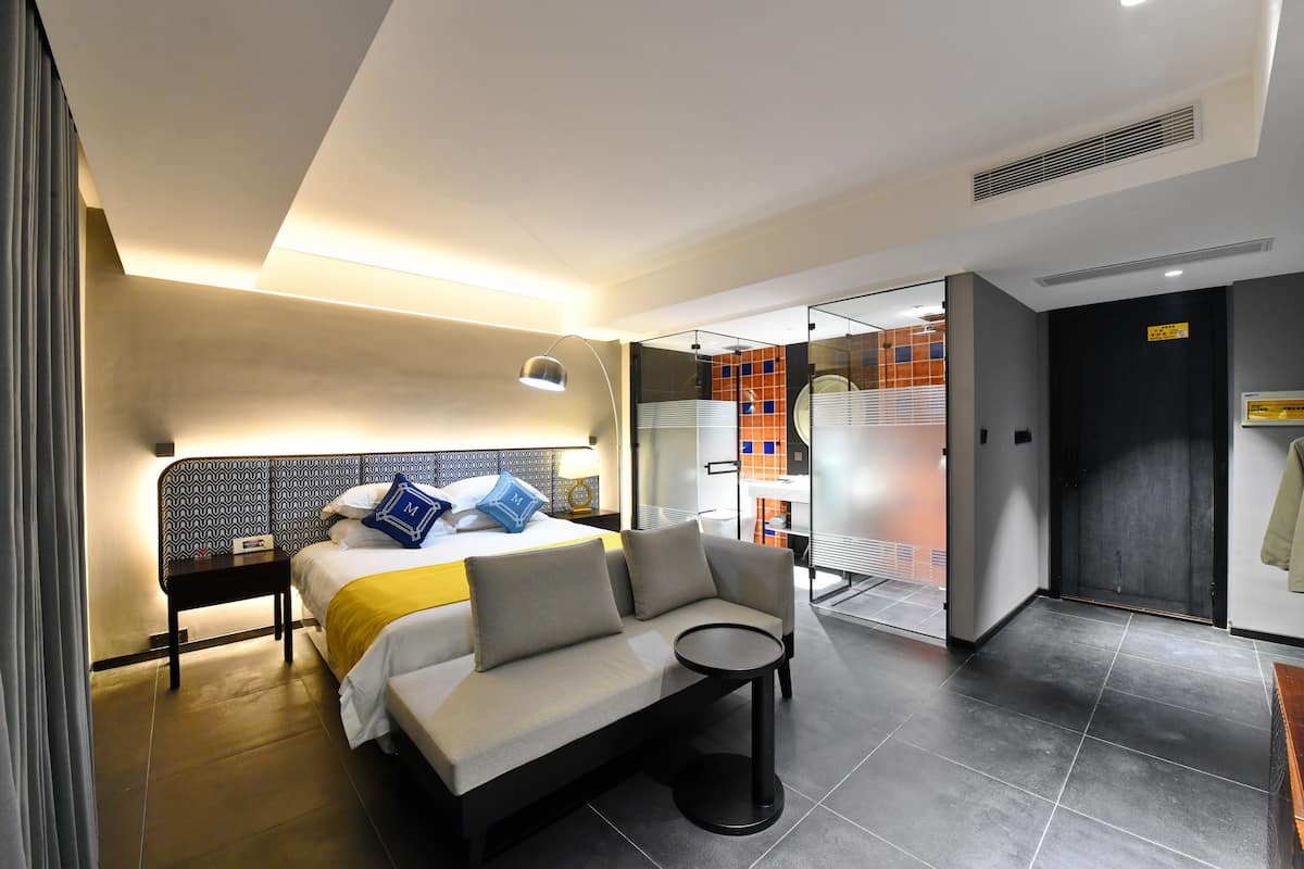 A modern hotel room with a bed, a sofa, a small table, and a glass partition separating the room from the hallway.