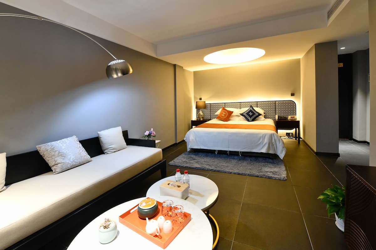 A modern hotel room with a sofa, a bed, a round coffee table, and a bedside table with a lamp.