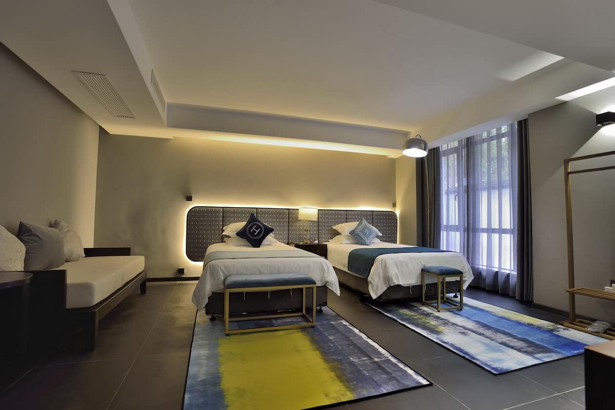 A modern hotel room with two beds, a sofa, a bench, and a large window with curtains.