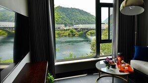 A modern hotel room with a large window offering a view of a river and a bridge, a sofa, a coffee table with a tray of items, and a hanging lamp. - Dreamhouse (Hangzhou)