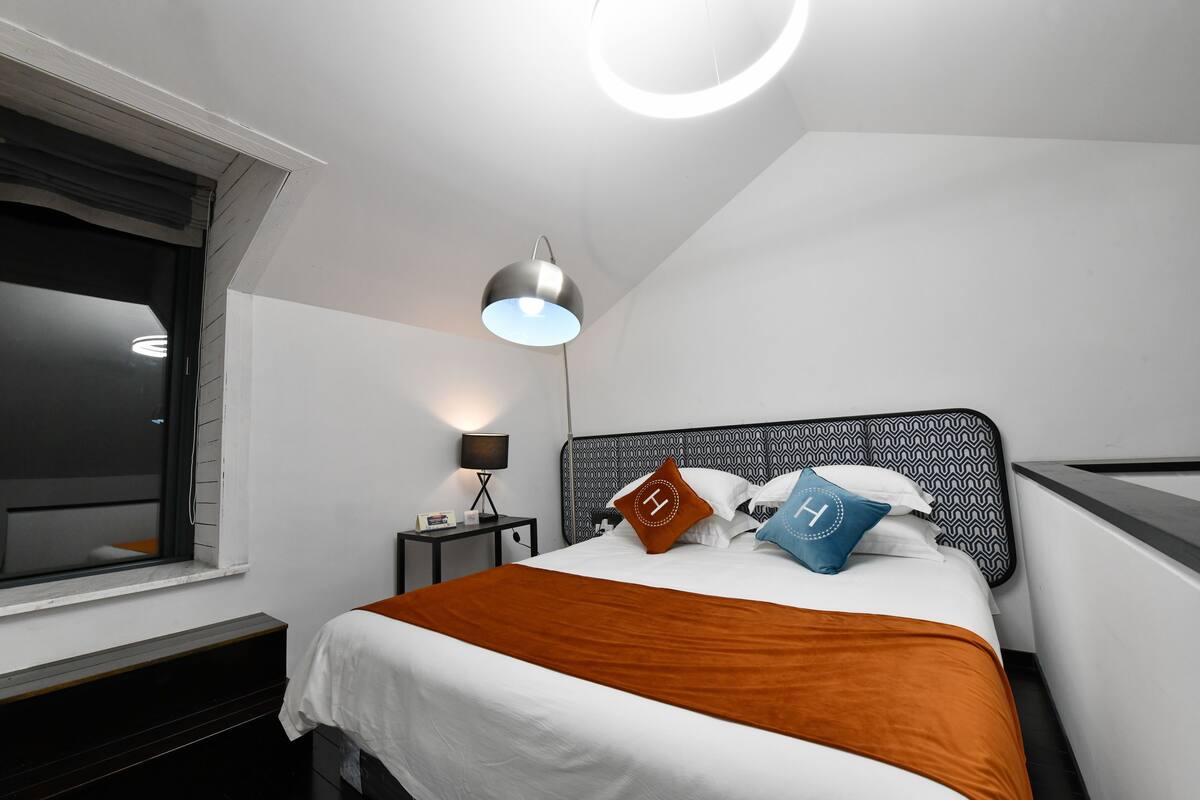 A modern hotel room with a neatly made bed, a bedside lamp, and a window with blinds.