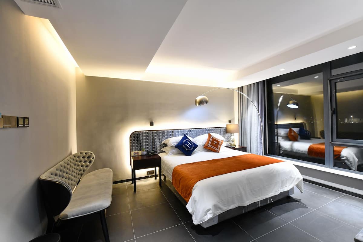 A modern hotel room with a large bed, a nightstand, a chair, and a large window offering a view of the city.