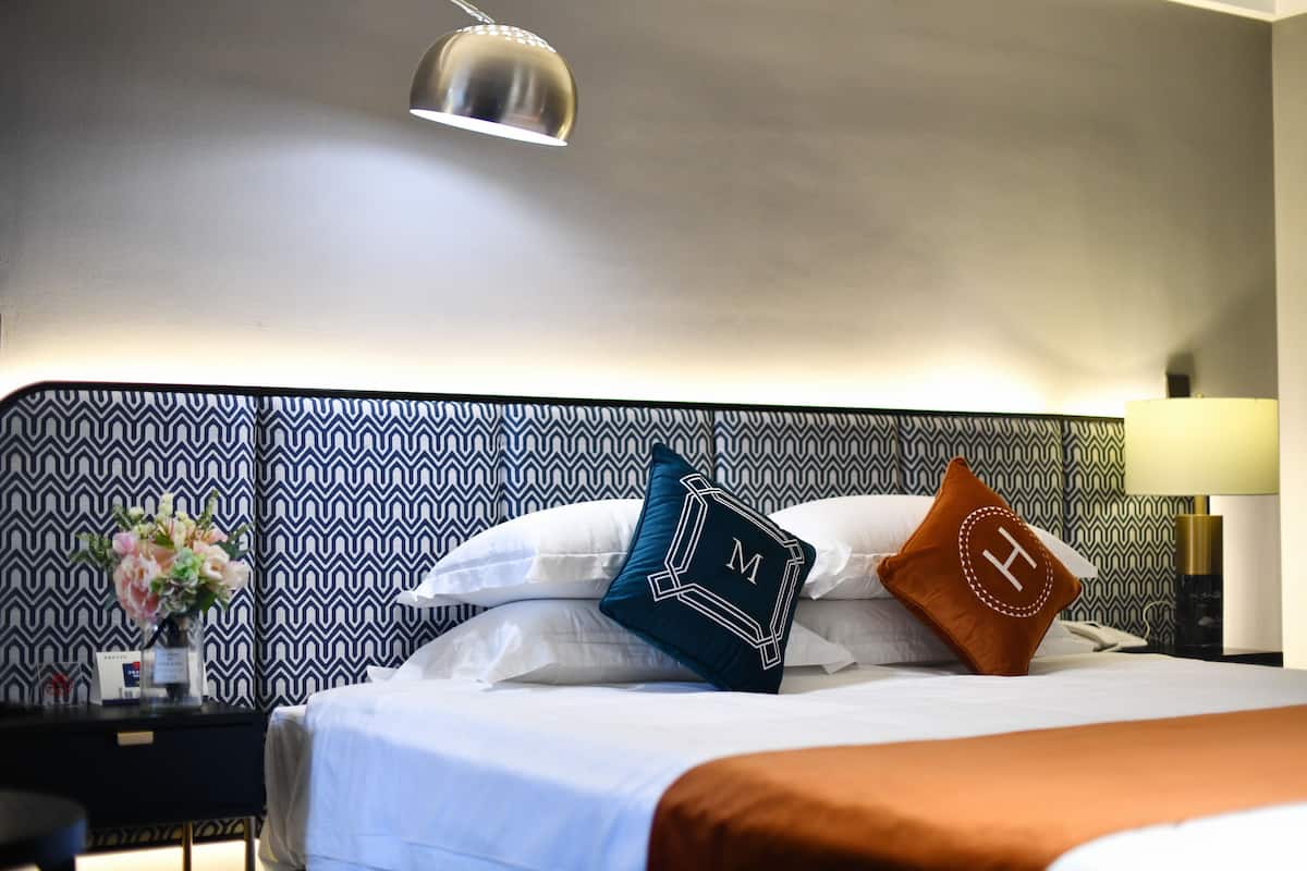 A hotel room with a patterned headboard, a bedside table with a lamp, and decorative pillows.