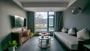 A modern living room with a sofa, a coffee table, a TV, and a large window with a view of a building and trees. - Dreamhouse (Hangzhou)