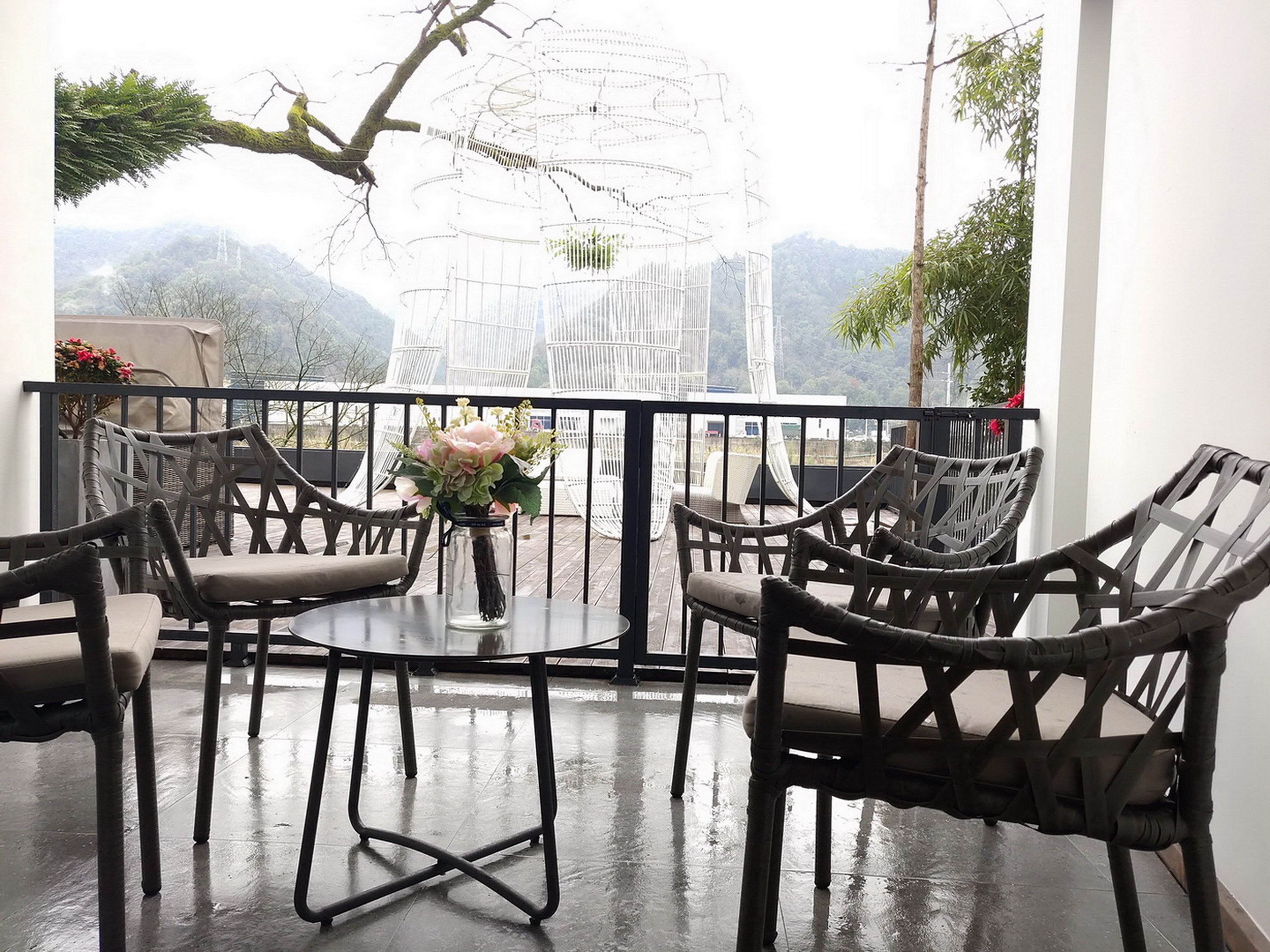 A balcony with wicker chairs and a table, a vase with flowers, and a view of mountains.