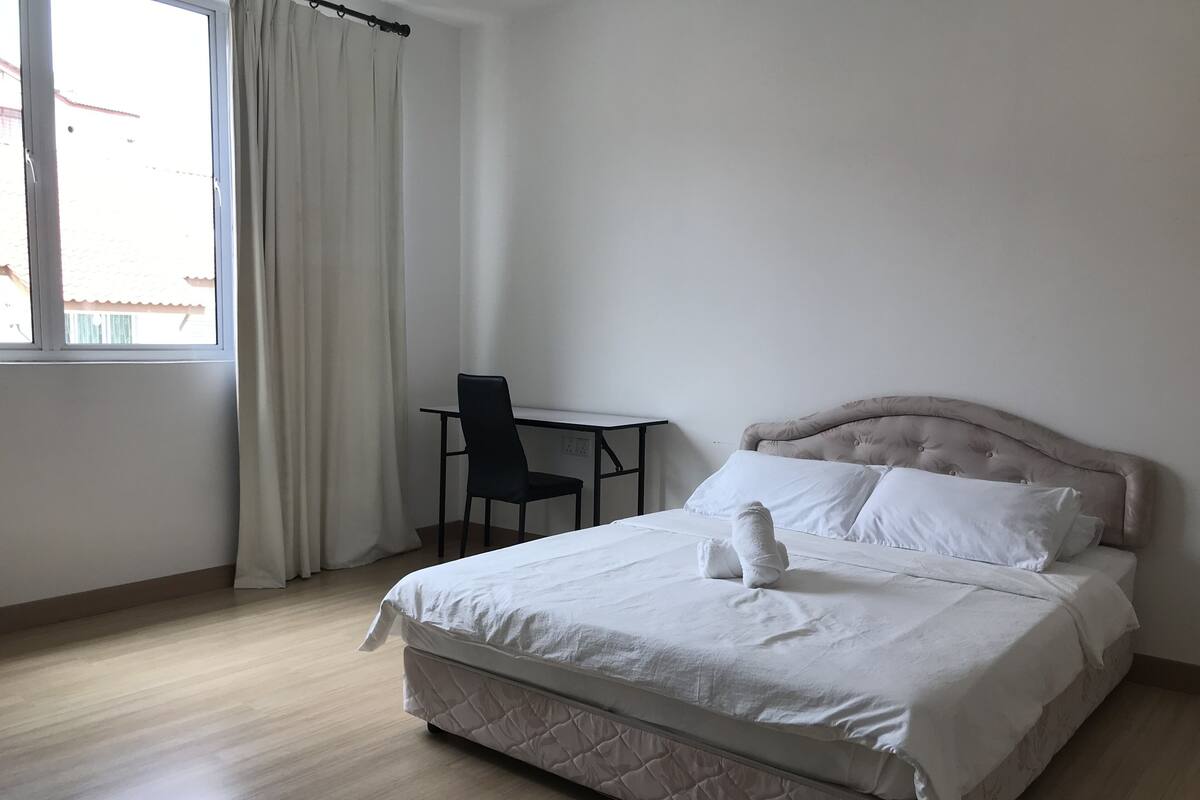 Un letto ben rifatto con materasso trapuntato e due cuscini, un tavolino con una sedia e una finestra con tende.
