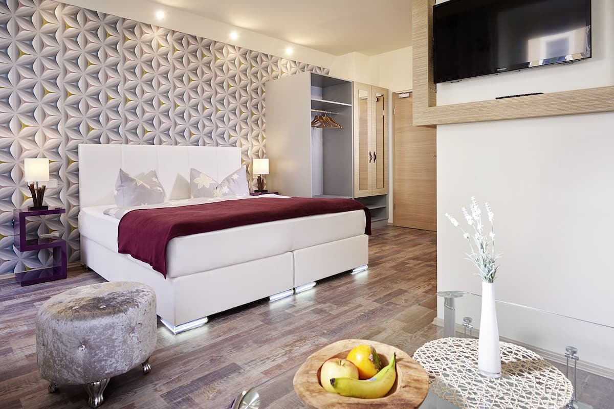 Une chambre moderne avec un mur à motifs, un lit recouvert d’une couverture rouge, une armoire, un téléviseur et un panier de fruits posé sur le sol.