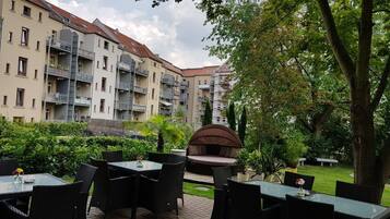 Ein Terrassenbereich im Freien mit Tischen und Stühlen, ein überdachter Sitzbereich und Blick auf mehrstöckige Gebäude mit Balkonen.