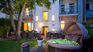 Ein Außenbereich mit Sitzgelegenheiten, darunter eine überdachte Couch, Esstische und Stühle, umgeben von Grünpflanzen und einem Gebäude mit Balkonen.