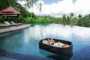 Uma piscina com uma bandeja flutuante de comida e bebidas, cadeiras de espreguiçadeira e uma estrutura com telhado de palha.