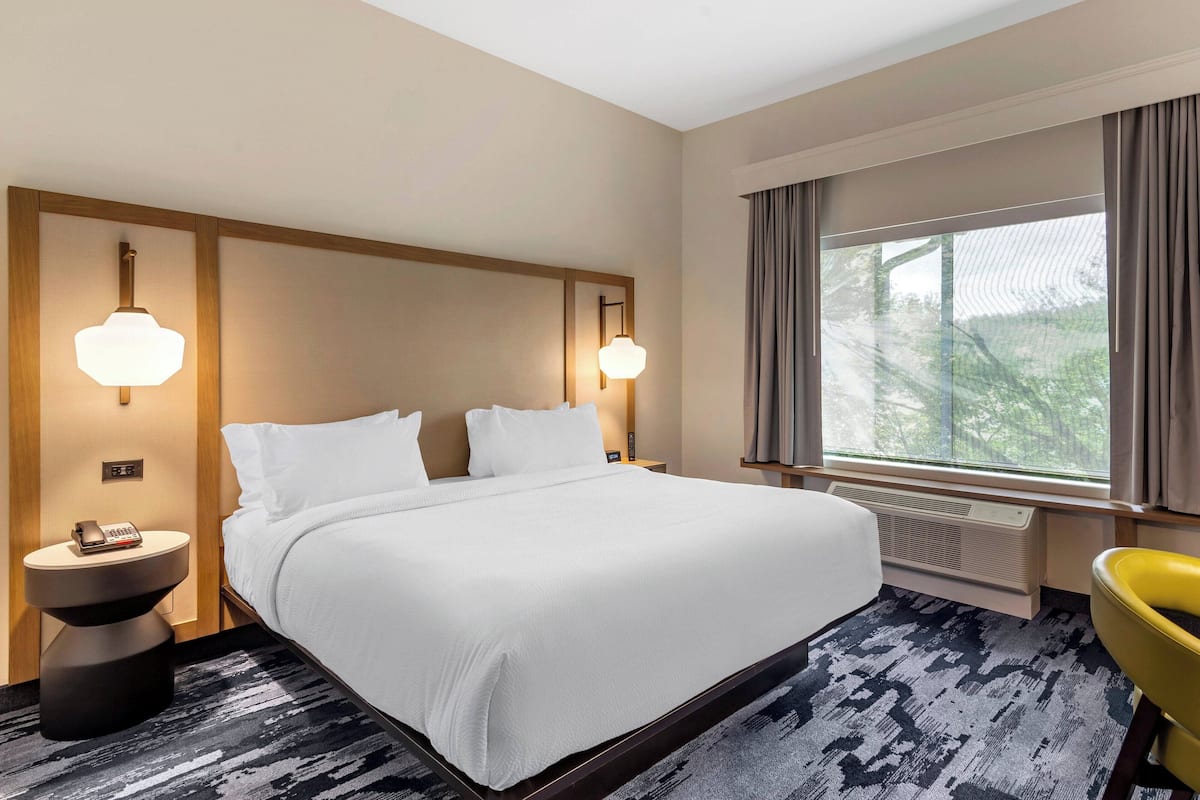 Une chambre d’hôtel avec un grand lit, deux lampes de chevet, un téléphone, un radiateur, une fenêtre avec des rideaux et une chaise jaune.