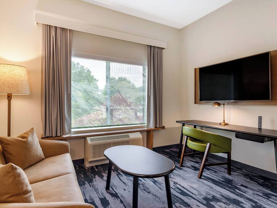 A hotel room with a sofa, a small table, a desk with a chair, a TV mounted on the wall, and a large window with curtains.