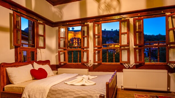 A bedroom with a large bed, wooden headboard, and multiple windows with curtains. - Beyzade Konak Otel (Safranbolu)