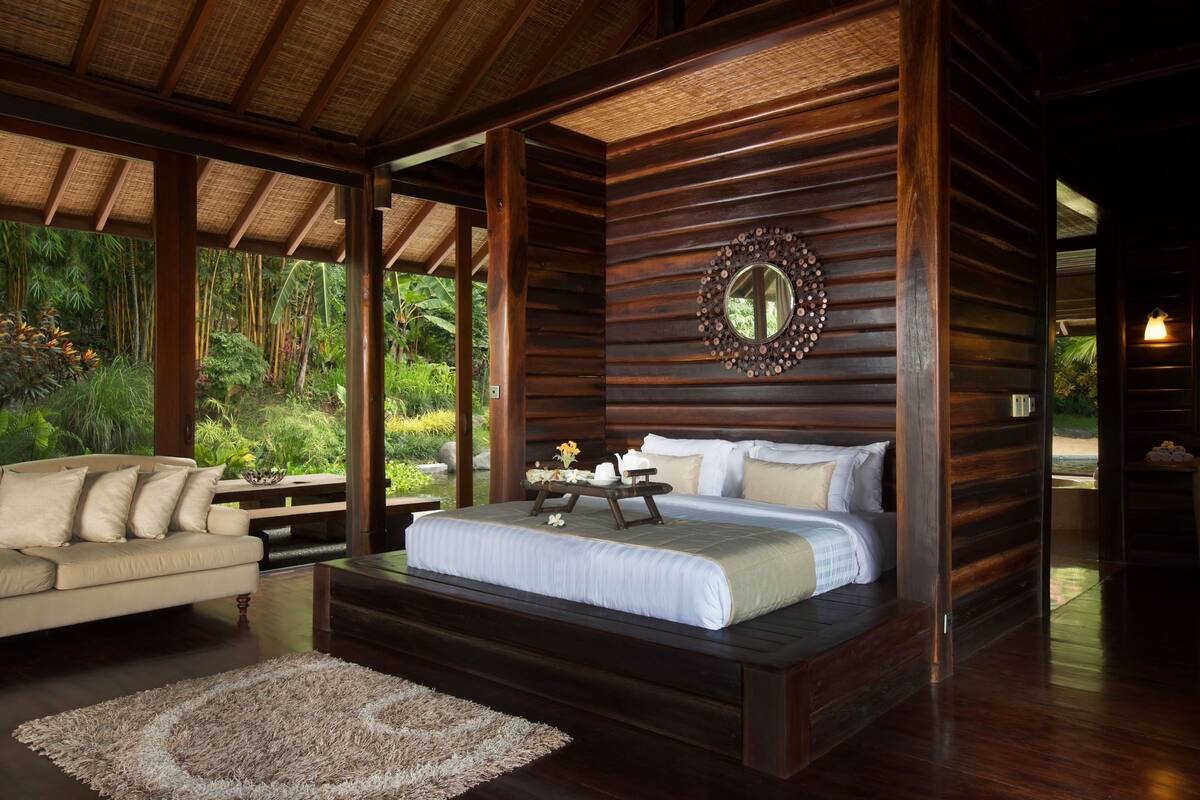 A wooden cabin-style bedroom with a bed, a sofa, and a view of lush greenery.