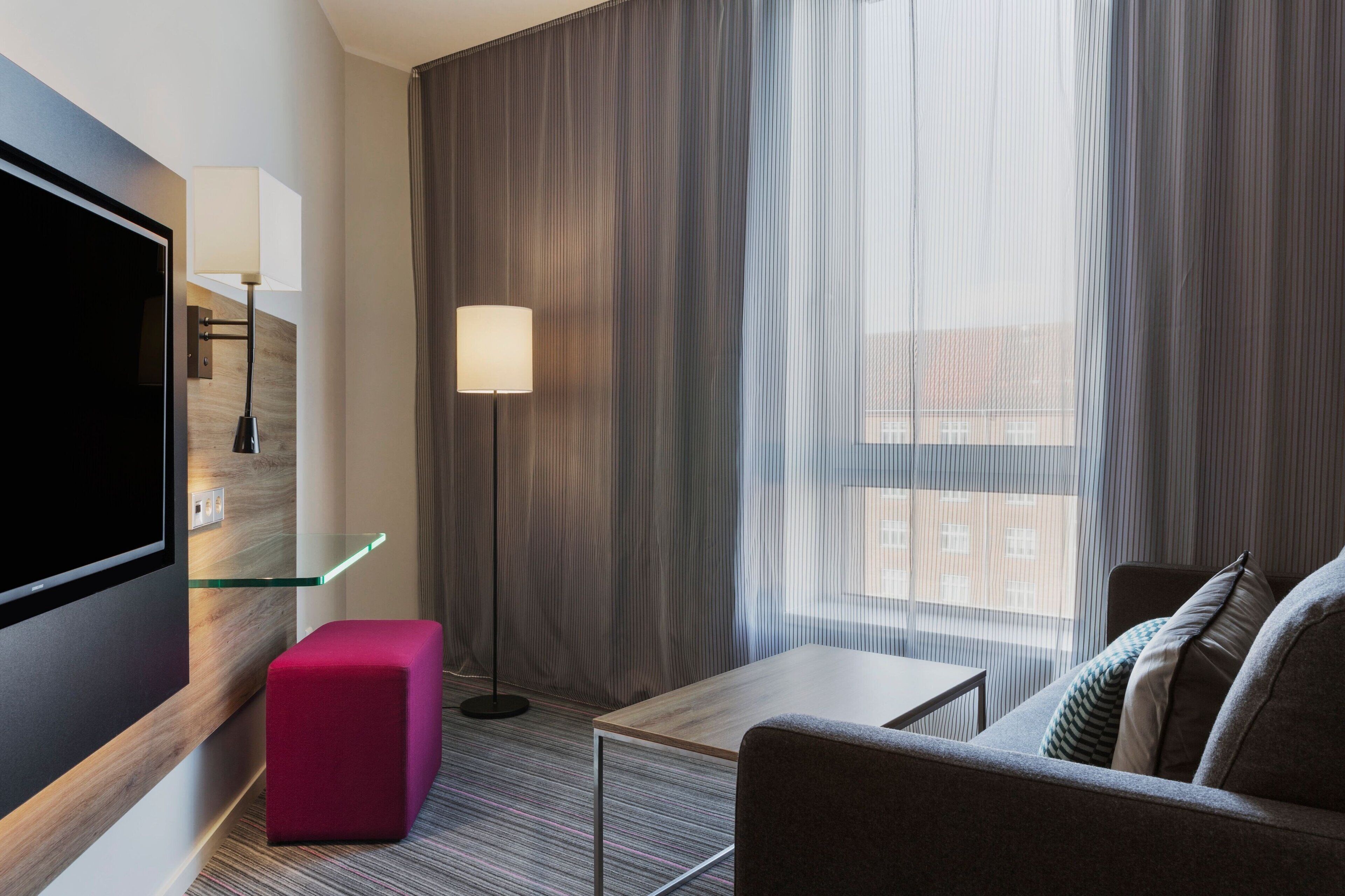 A modern hotel room with a flat-screen TV, a glass shelf, a red ottoman, a grey sofa, a small table, a floor lamp, and a large window with curtains.