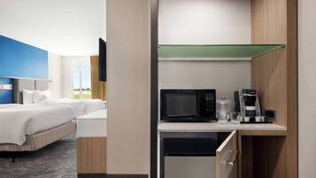 A compact hotel room with a bed, a small kitchenette with a microwave, coffee maker, and glass, and a view of the outdoors through a window.