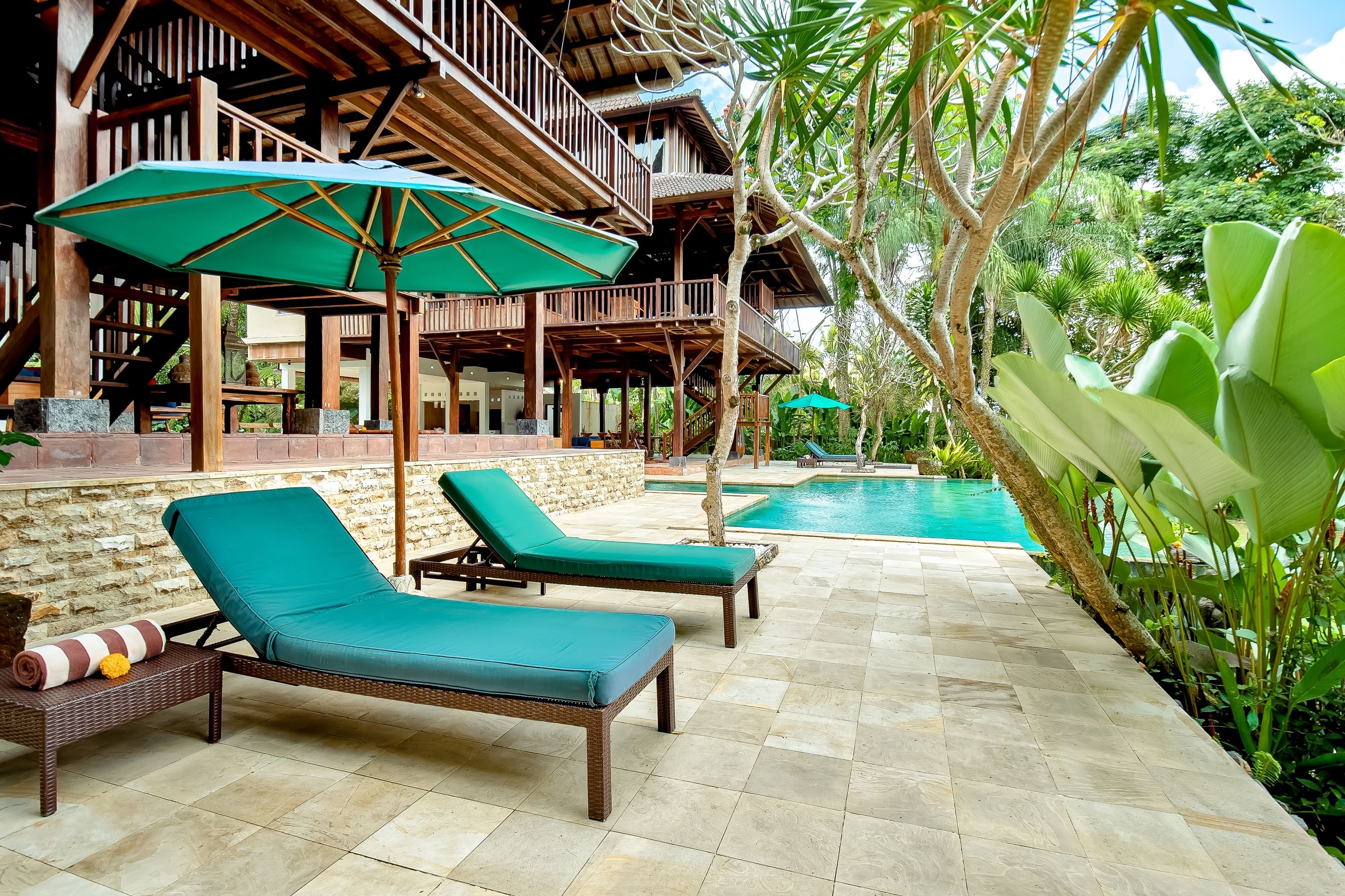 A poolside area with lounge chairs and a large umbrella, surrounded by lush greenery and a wooden structure.