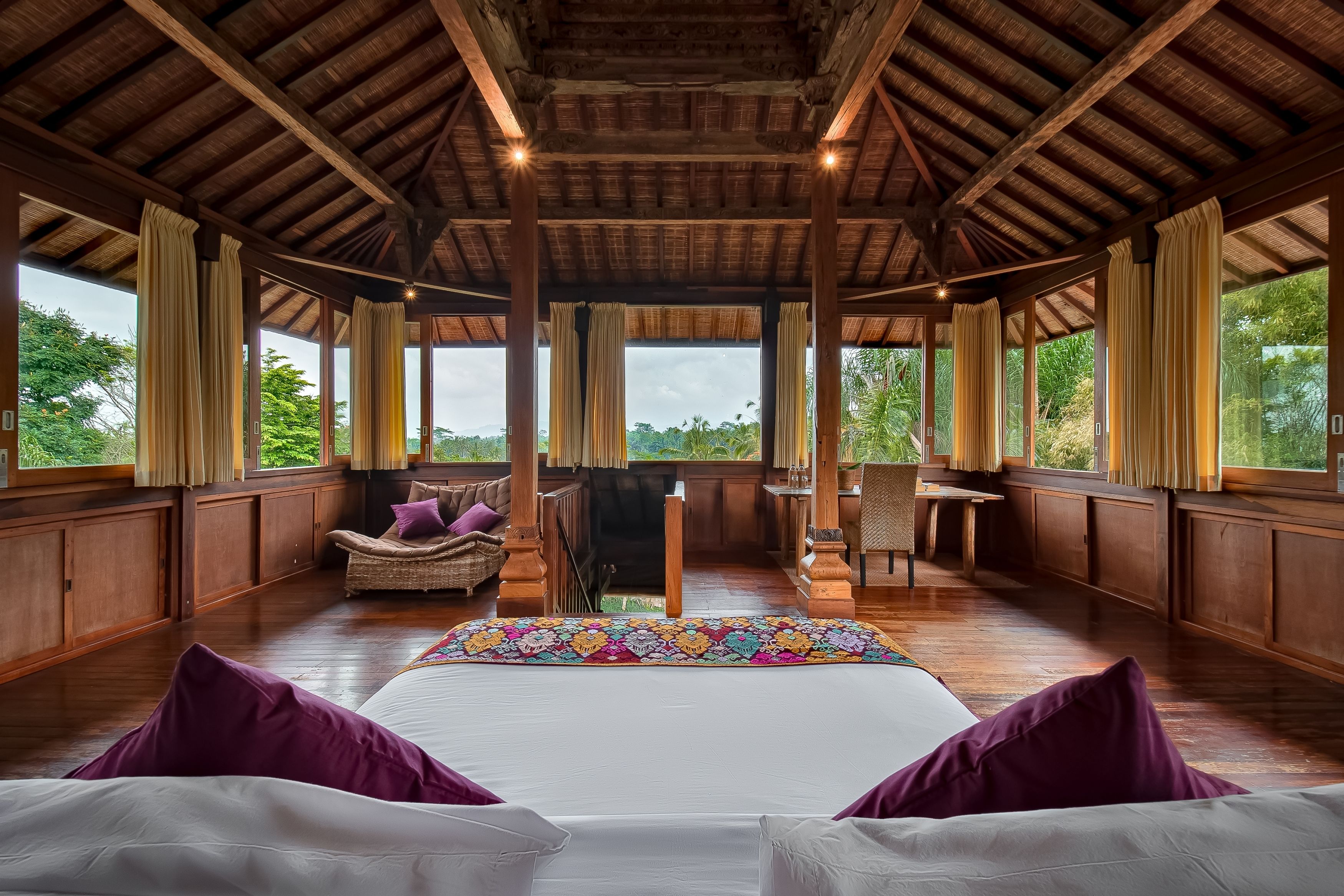 A spacious room with a large bed, wooden ceiling, and large windows offering a view of greenery.