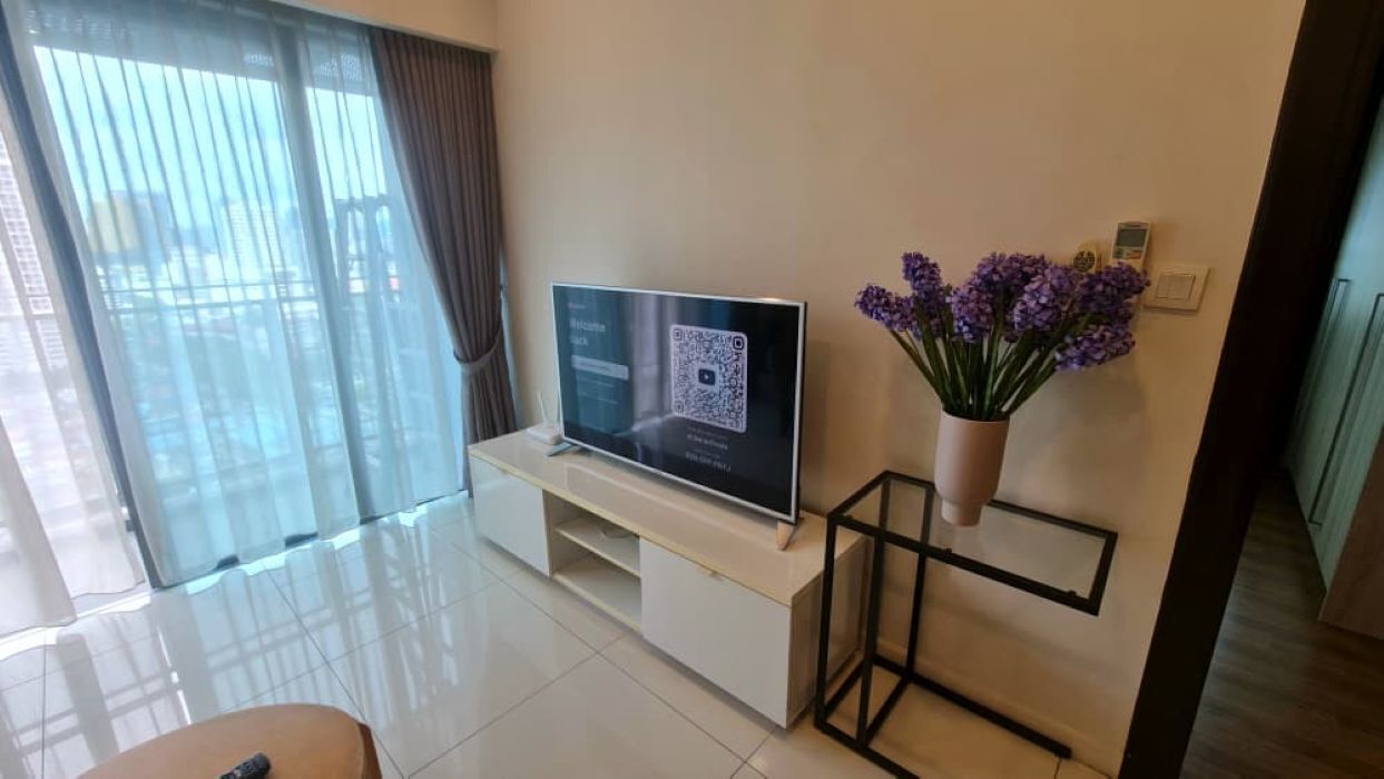 A modern living room with a flat-screen TV on a white entertainment unit, a small table with a vase of purple flowers, and a sliding glass door leading to a balcony.