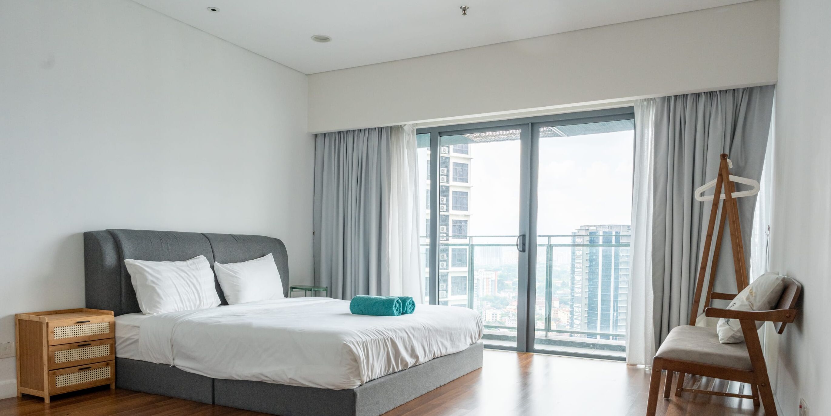 A modern bedroom with a large bed, a wooden nightstand, a chair, and a view of the city through large windows.