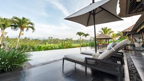 A modern outdoor lounge area with a large umbrella, lounge chairs, and a view of a lush landscape. - Villa Rumah Lotus (Payangan)