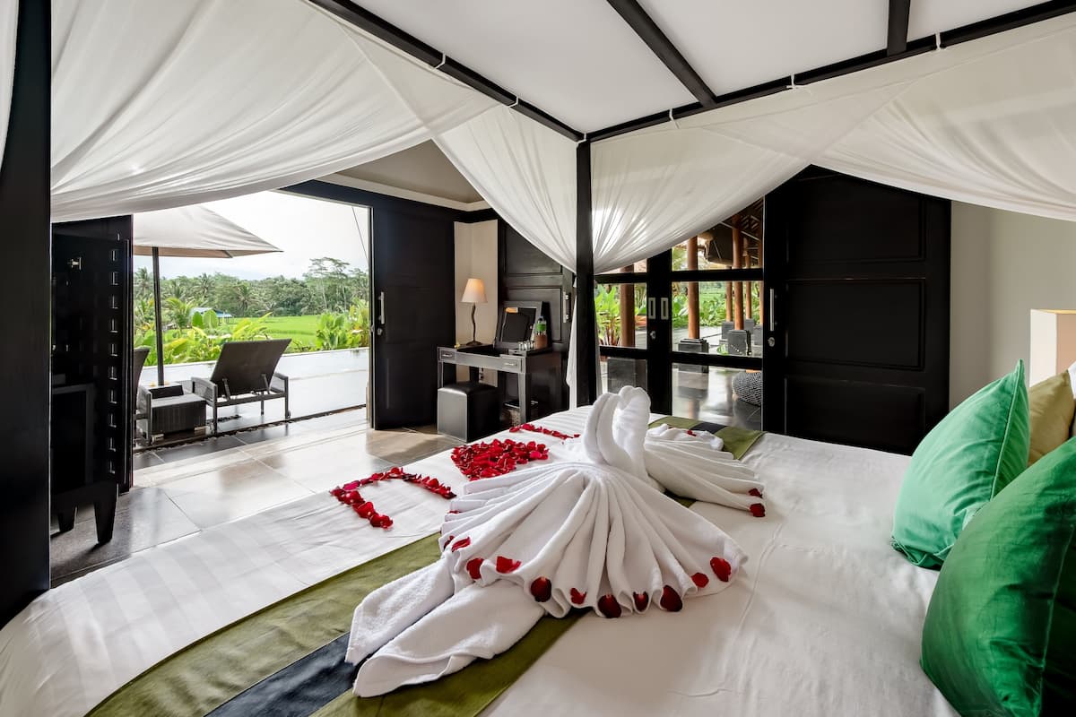 A bedroom with a bed covered in white linens and red rose petals, a green pillow, and a canopy.
