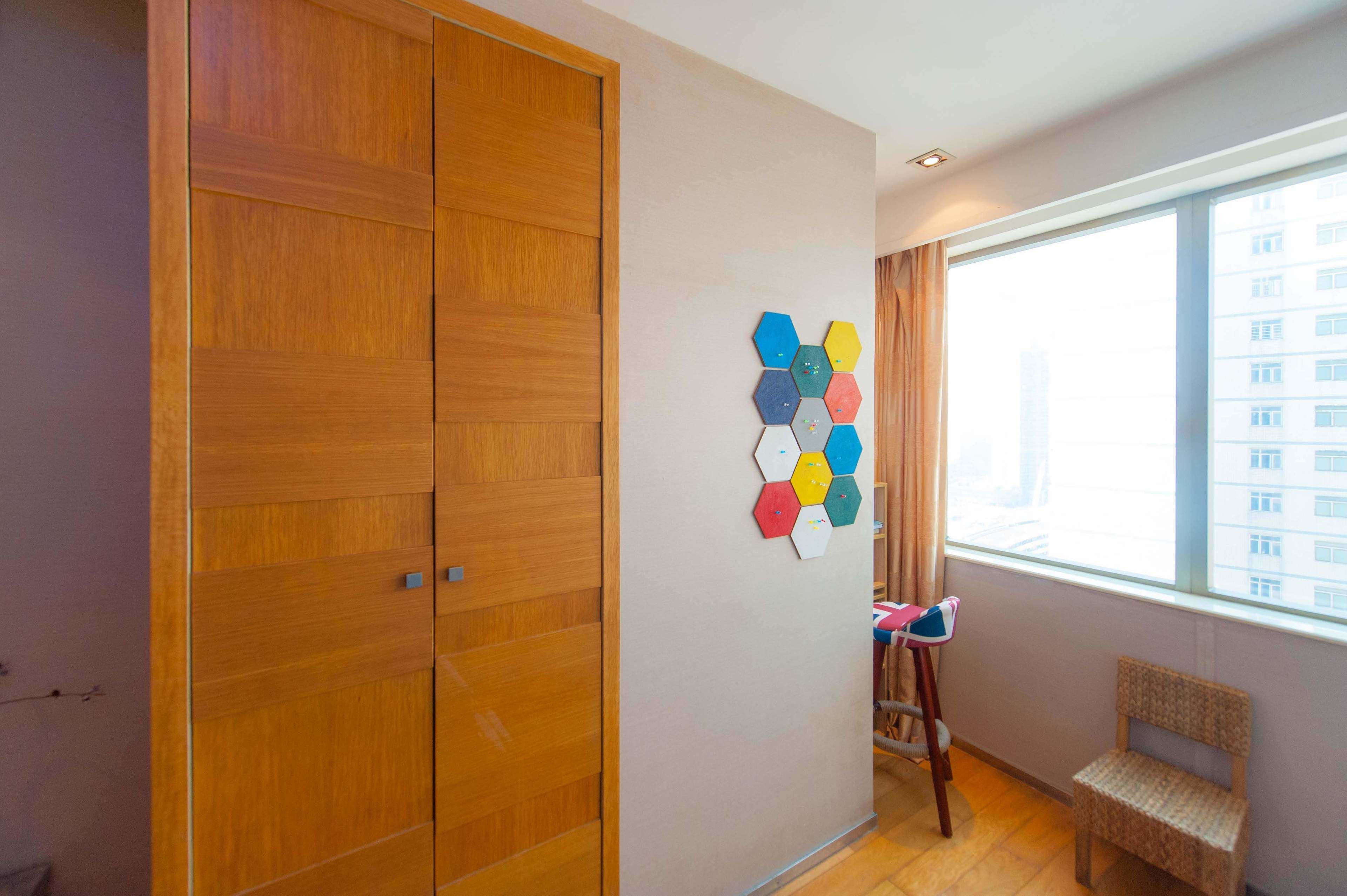 Habitació amb un armari de fusta, una decoració mural hexagonal i colorida, una cadira petita i una finestra que dona a edificis.