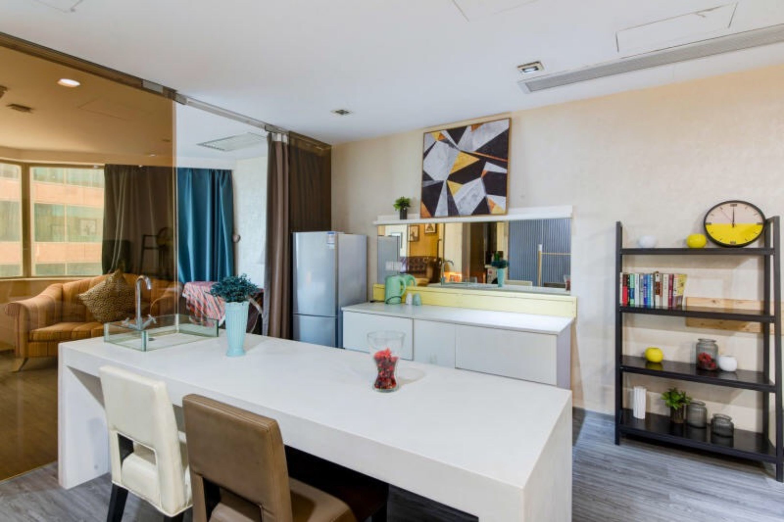 A modern hotel room with a kitchenette, a dining area with a white table and chairs, a sofa, a bookshelf, and a large abstract painting on the wall.