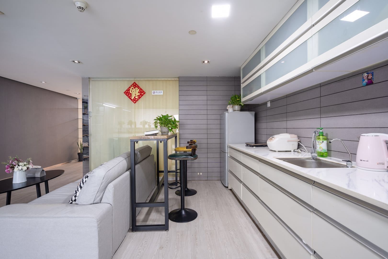 A modern kitchen with a white countertop, a sink, and a refrigerator. There is a small table with a plant and a vase of flowers.