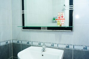 A bathroom with a glass shelf above a white sink, tiled walls, and a mirror reflecting a toilet paper dispenser and a soap dispenser. - Hotel Meigah (Tanjung Pandan)