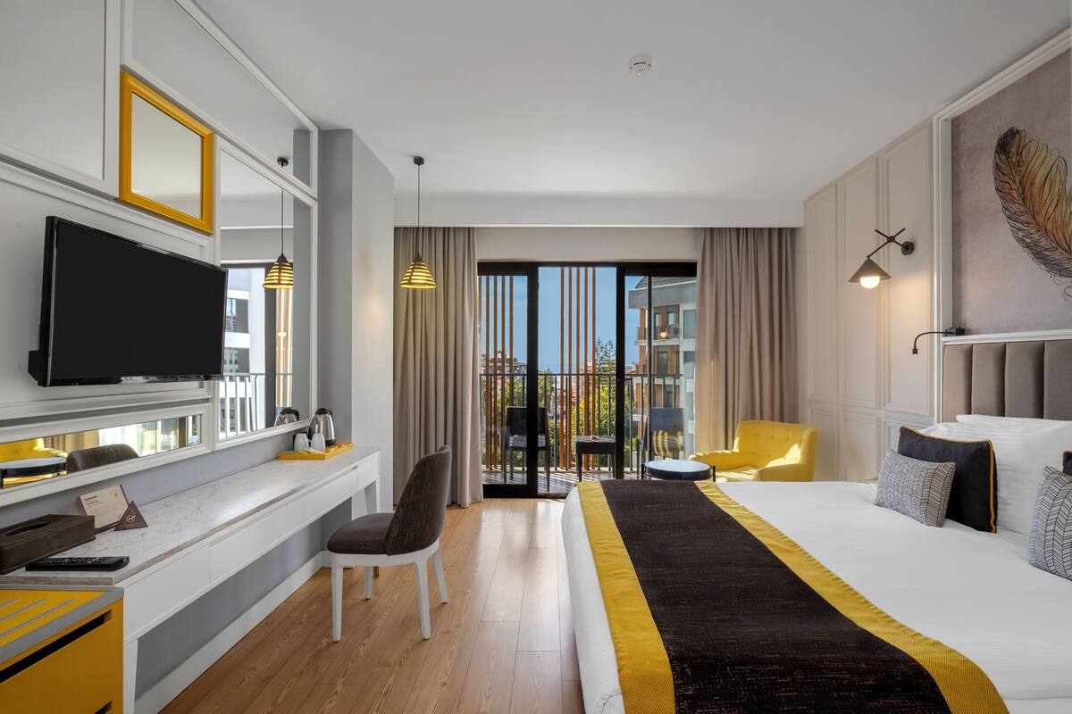 Habitación de hotel con una cama grande, un escritorio con televisión de pantalla plana, una silla, un sofá y un balcón con vista a edificios.