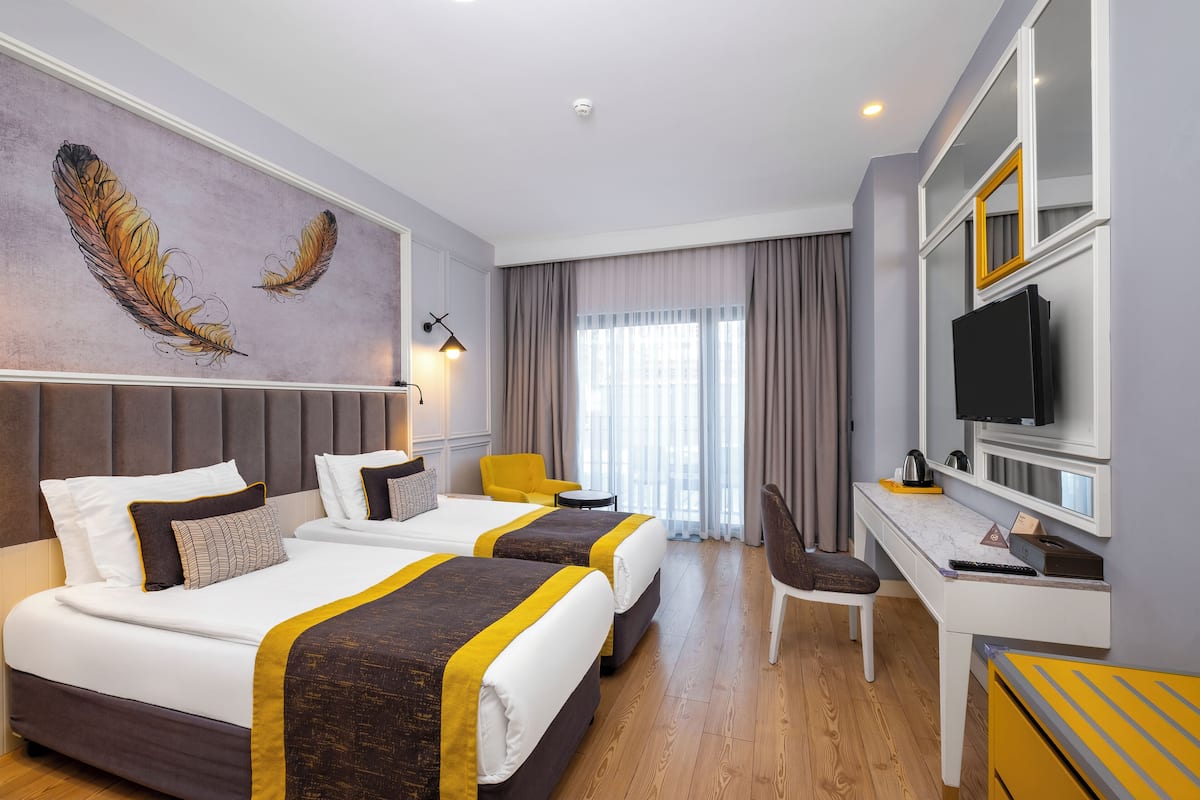 Habitación de hotel con dos camas, un escritorio, una silla, un televisor y una gran obra de arte de plumas en la pared.